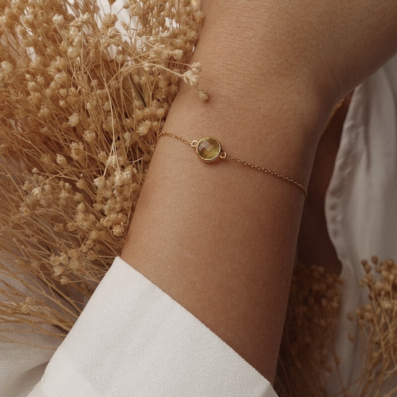 Close-up of a delicate gold chain bracelet with a round green gemstone on a wrist against dried flowers background