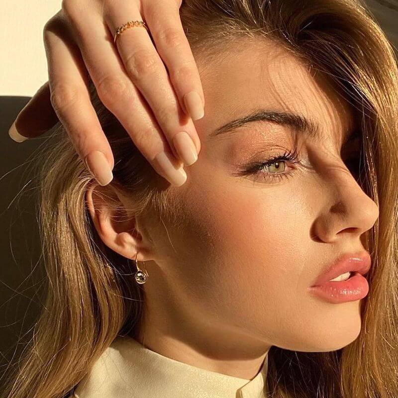 Close-up of a woman with light makeup and natural nails resting her hand on her forehead in warm sunlight