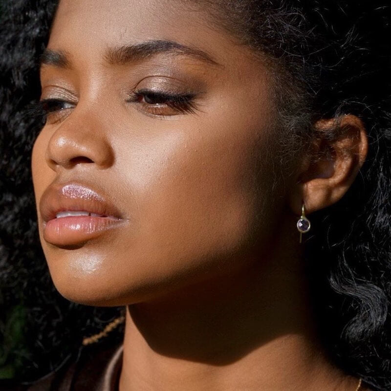 Close-up of a woman with natural makeup and curly hair wearing a small round drop earring against a dark background