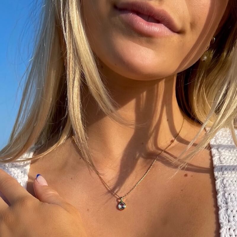Close-up of a woman with blonde hair wearing a delicate gold necklace with a small pendant against a clear blue sky