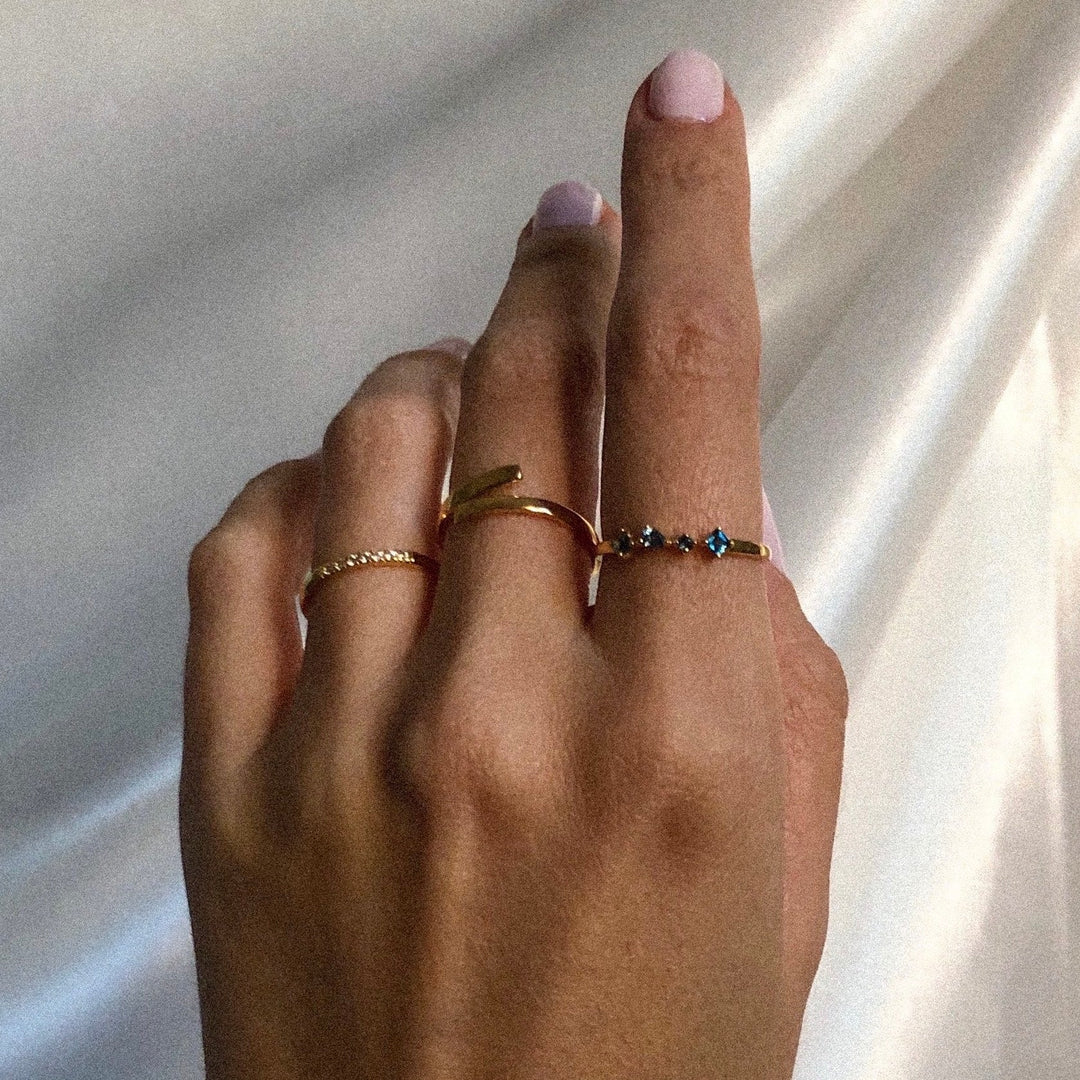 Close-up of a hand with light pink nail polish wearing three delicate gold rings against a soft white fabric background