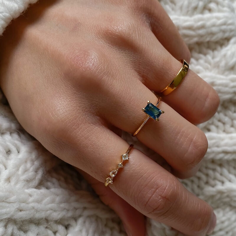 Close-up of a hand wearing three delicate gold rings, one with a rectangular blue gemstone, resting on a knitted cream fabric