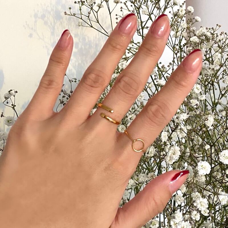 Close-up of a hand with glossy red-tipped nude nails wearing two delicate gold rings against white flowers background