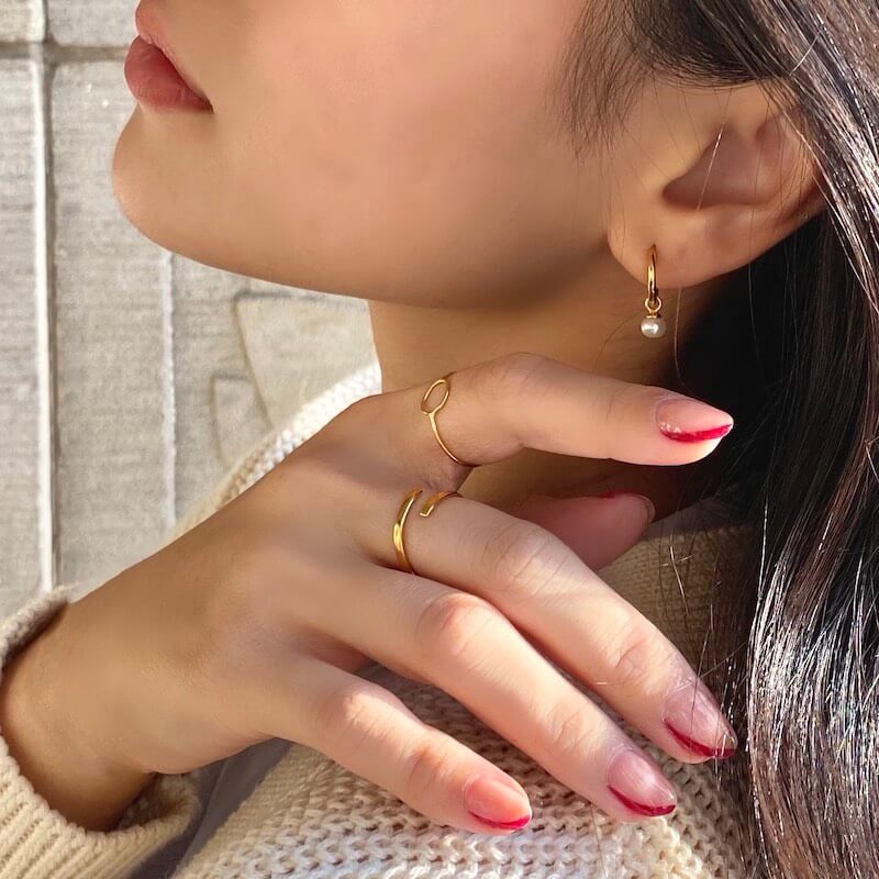 Close-up of a woman’s hand with gold rings and red-tipped nails gently touching her neck wearing a gold hoop earring with a pearl pendant
