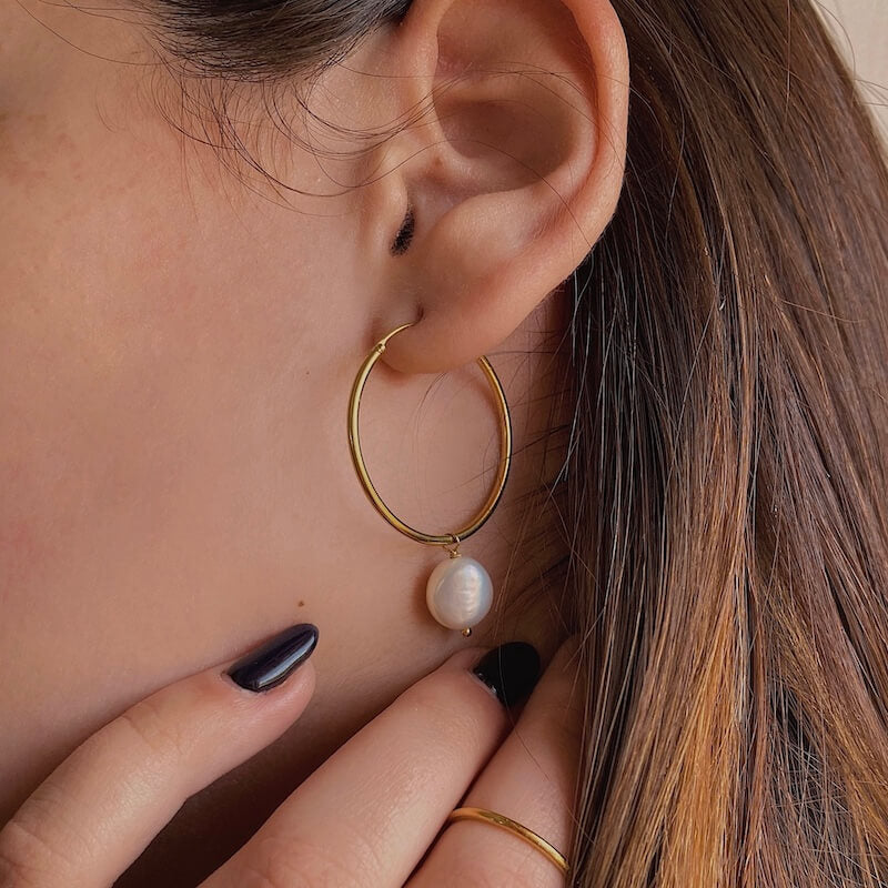 Close-up of a gold hoop earring with a single white pearl worn by a person with dark brown hair and black nail polish