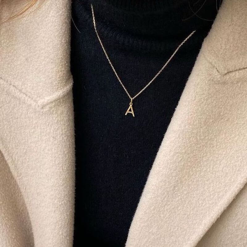 Close-up of a gold initial A pendant necklace worn over a black turtleneck and beige wool coat