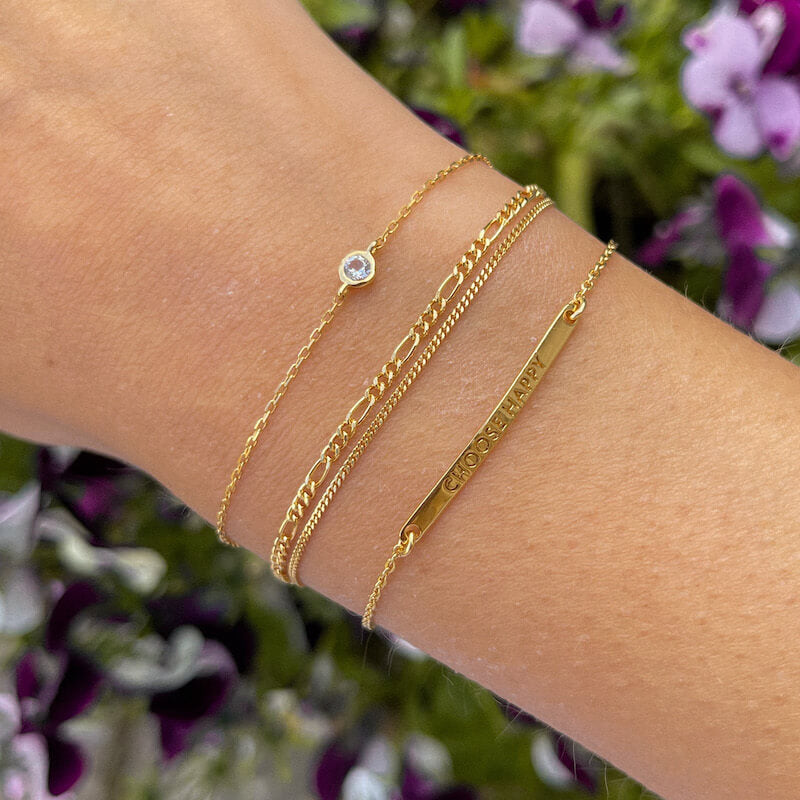 Close-up of a wrist wearing three delicate gold chain bracelets with one featuring a small clear stone and a rectangular bar charm outdoors