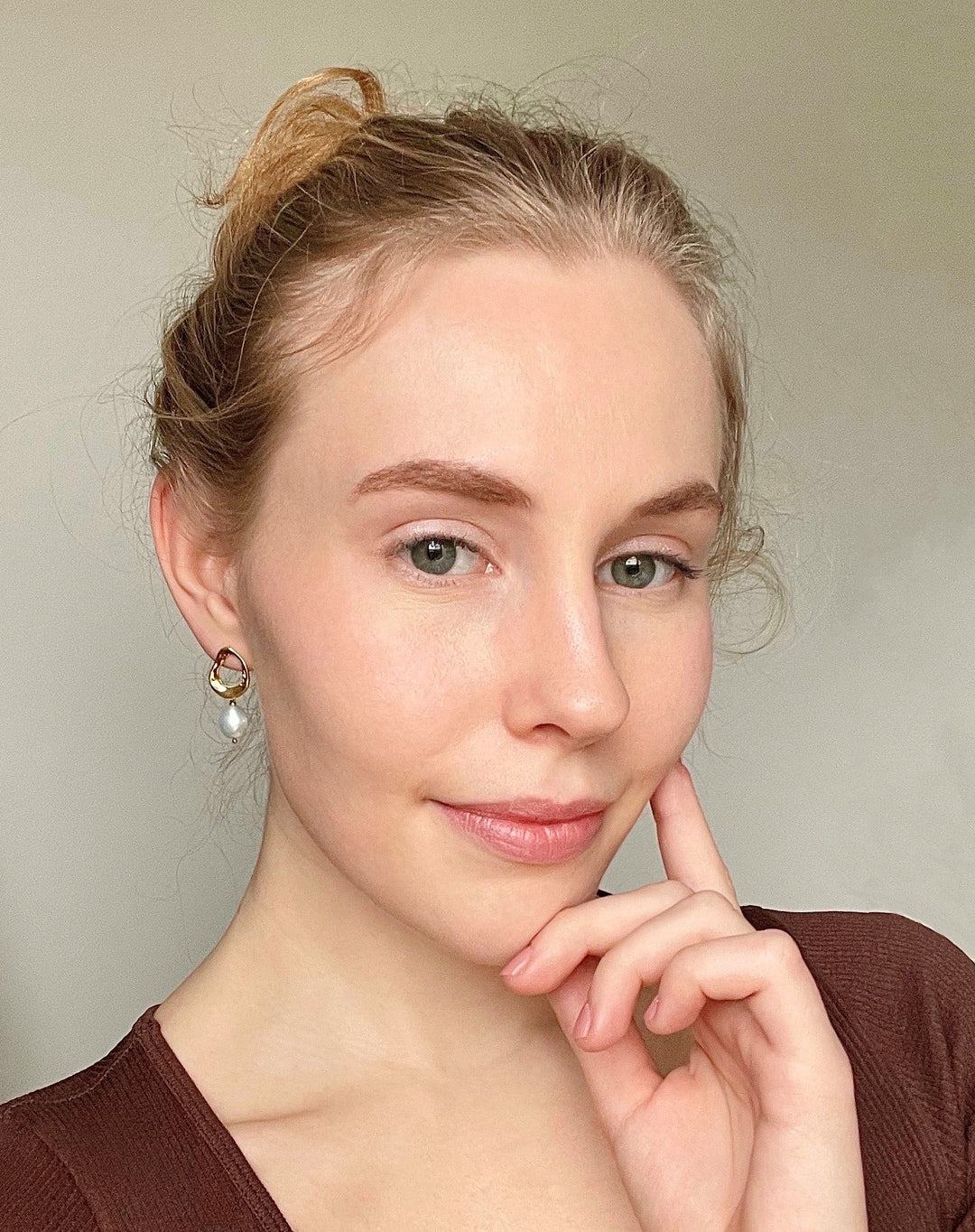 Close-up of a woman with light makeup wearing gold hoop earrings with dangling pearls against a plain beige background