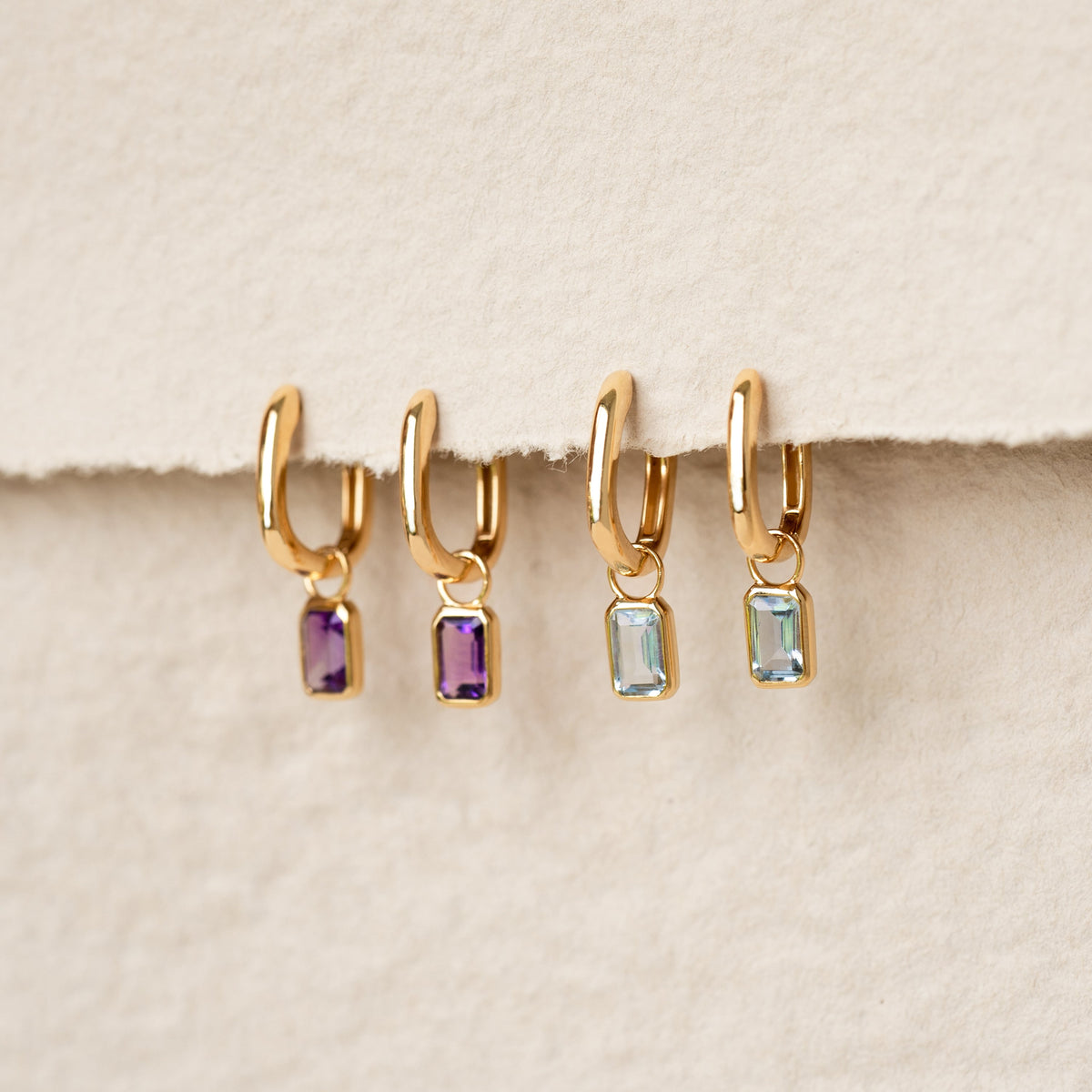Four gold vermeil Huggie Earrings with Baguette Charms Amethyst - Amelia in purple and light blue stones on a beige background
