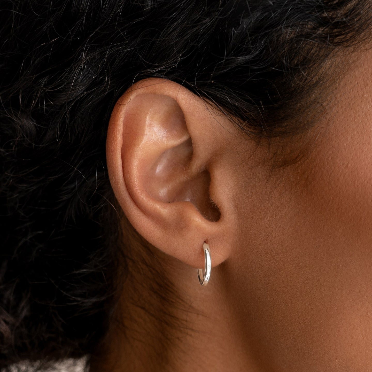 Close-up of an ear wearing White Gold Huggie Earrings - Emilia with a smooth hinge closure on a neutral background