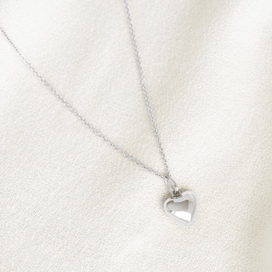 Close-up of a Puffy Heart Necklace Silver with a shiny heart pendant on a delicate silver chain against a cream fabric background