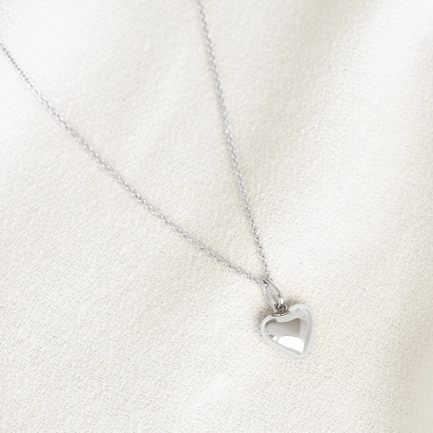 Close-up of a Puffy Heart Necklace Silver with a shiny heart pendant on a delicate silver chain against a cream fabric background