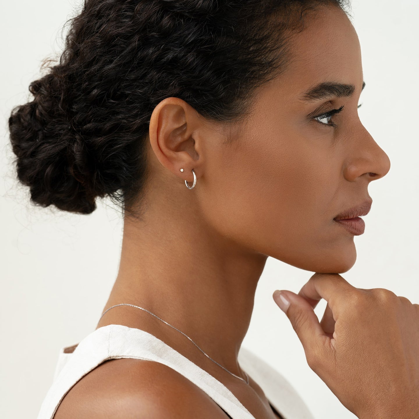 Profile of a woman wearing White Gold Huggie Earrings - Emilia and a delicate necklace against a light background
