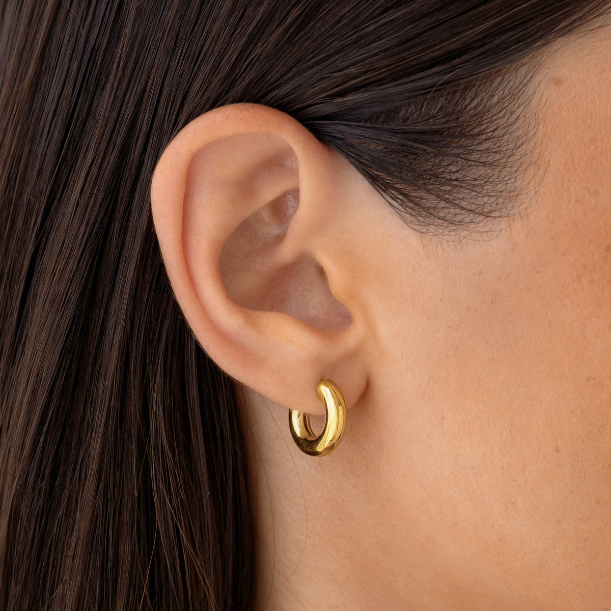 Close-up of a gold Chunky Huggie Hoop Earrings - Tove worn on a woman's ear with smooth skin and dark hair