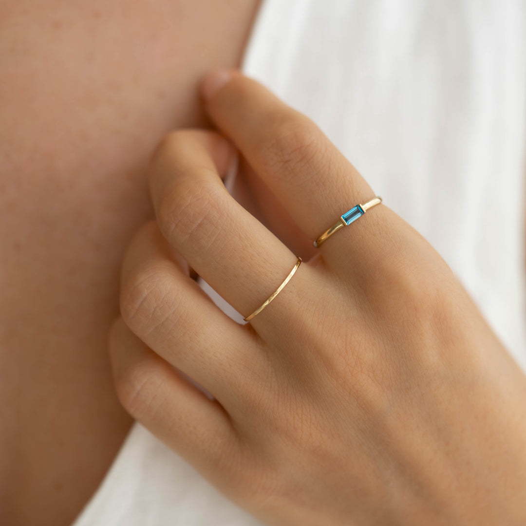 Close-up of hand wearing Baguette Ring London Blue Topaz 14k Gold - Synne with a slim gold band on a light background