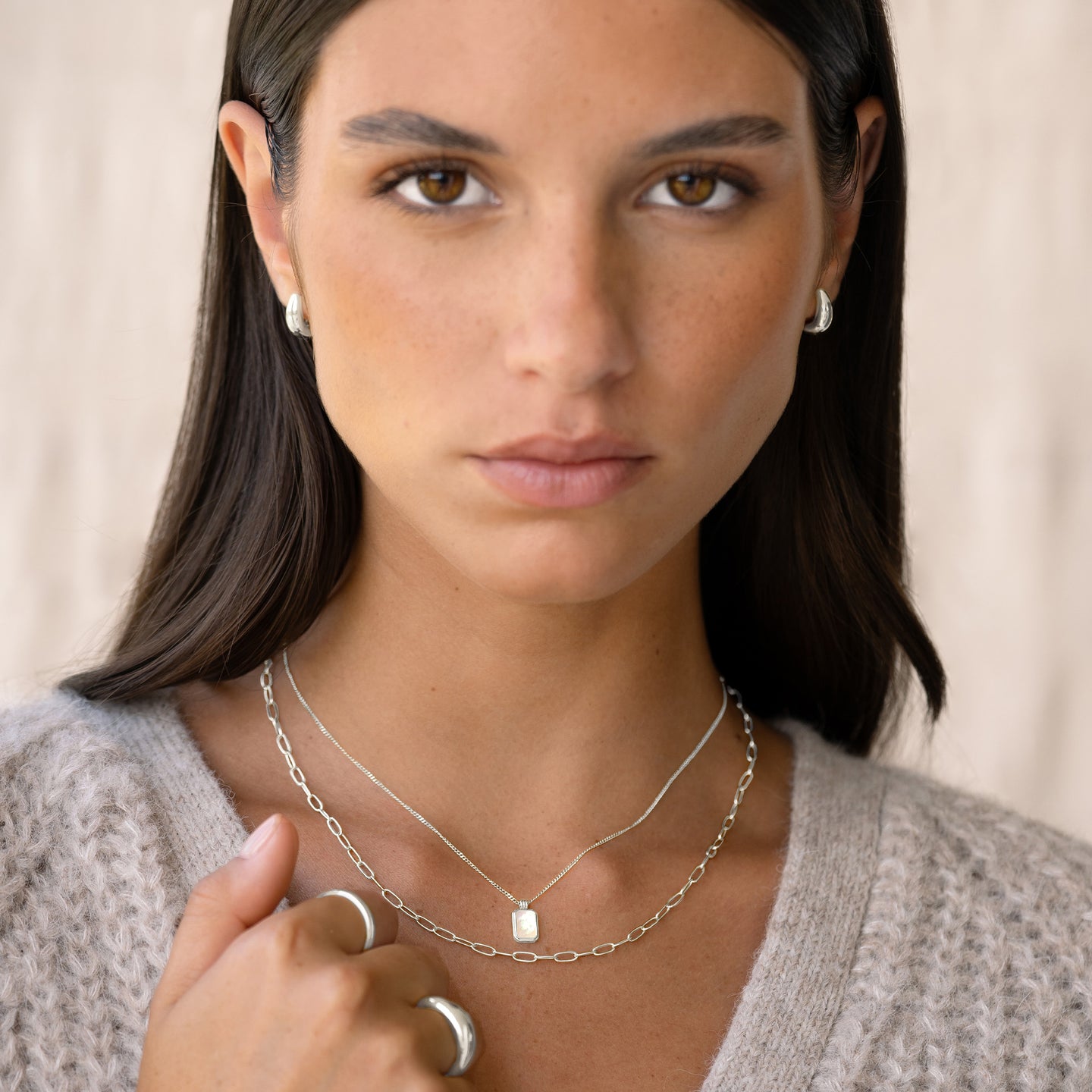 Close-up of a woman wearing Dome Huggie Earrings Silver - Caja with layered silver necklaces and rings on a neutral background