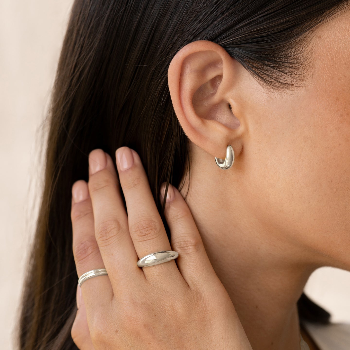 Close-up of a woman wearing Dome Huggie Earrings Silver - Caja and silver rings with dark hair and neutral background