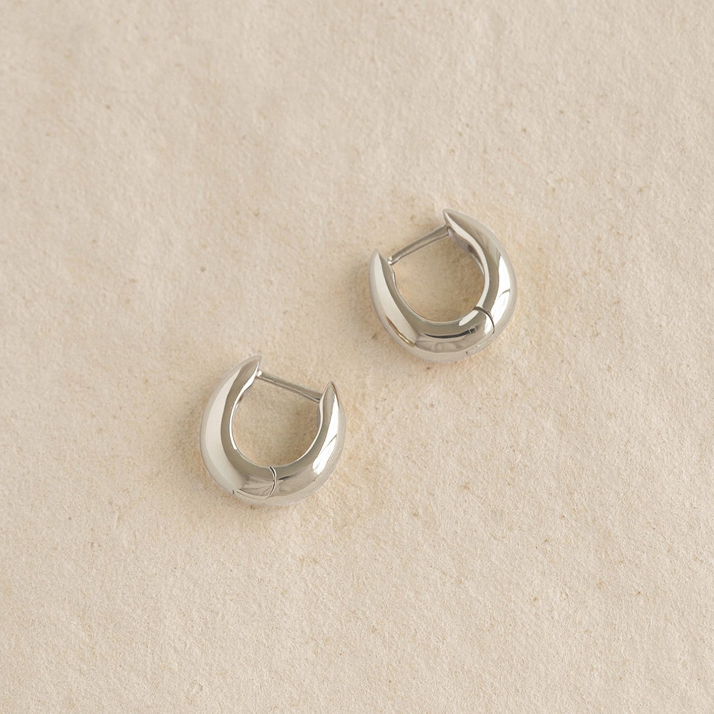 Pair of Dome Huggie Earrings Silver - Caja with subtle tapered dome shape on a beige textured surface