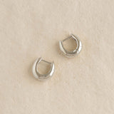Pair of Dome Huggie Earrings Silver - Caja with subtle tapered dome shape on a beige textured surface
