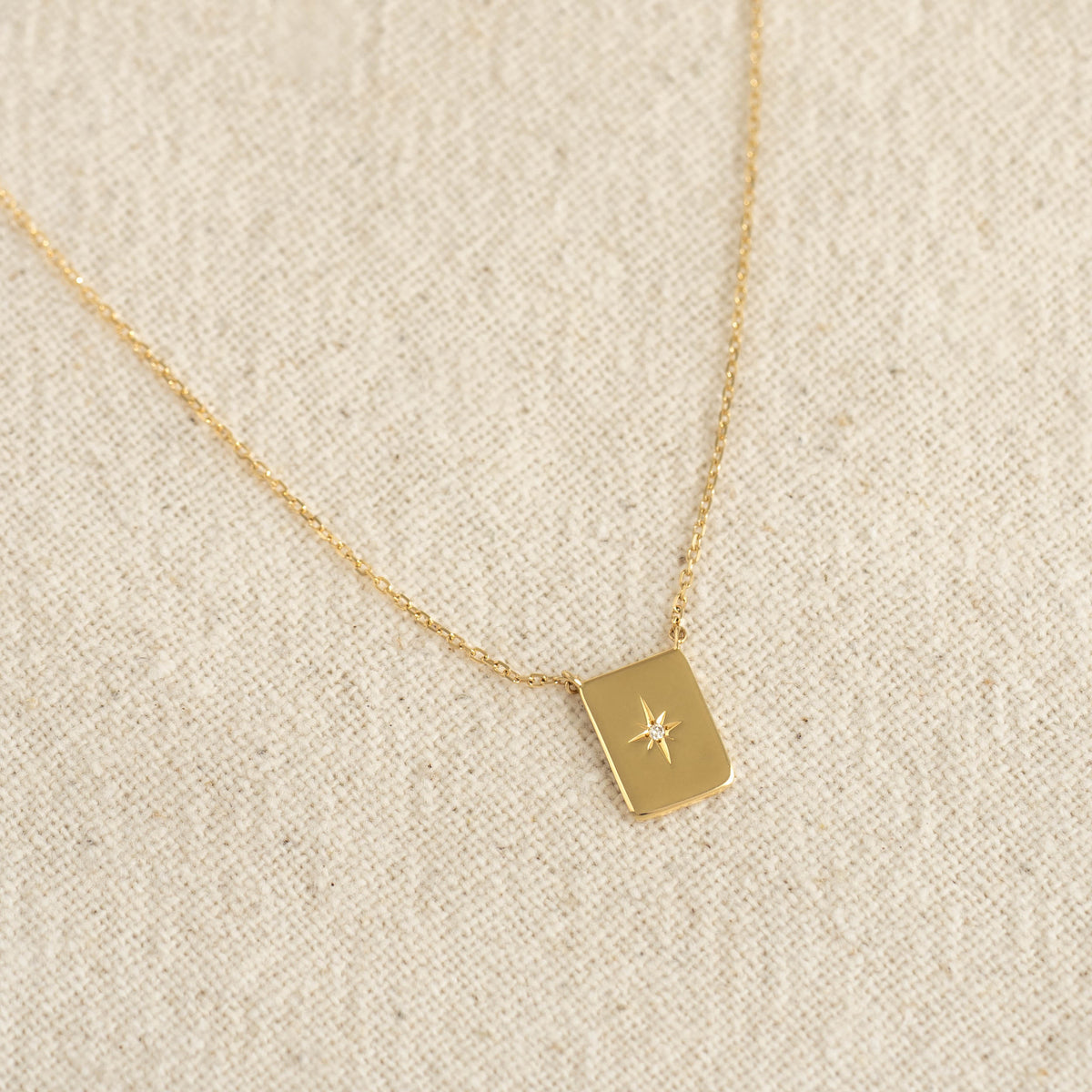 Close-up of Diamond Star Necklace - Polaris with gold tag pendant featuring star engraving and central diamond on textured fabric background