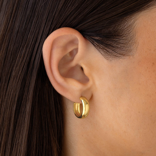 Close-up of ear wearing Small Bold Hoop Earrings - Ebba in gold vermeil with a wide hoop design on a neutral background