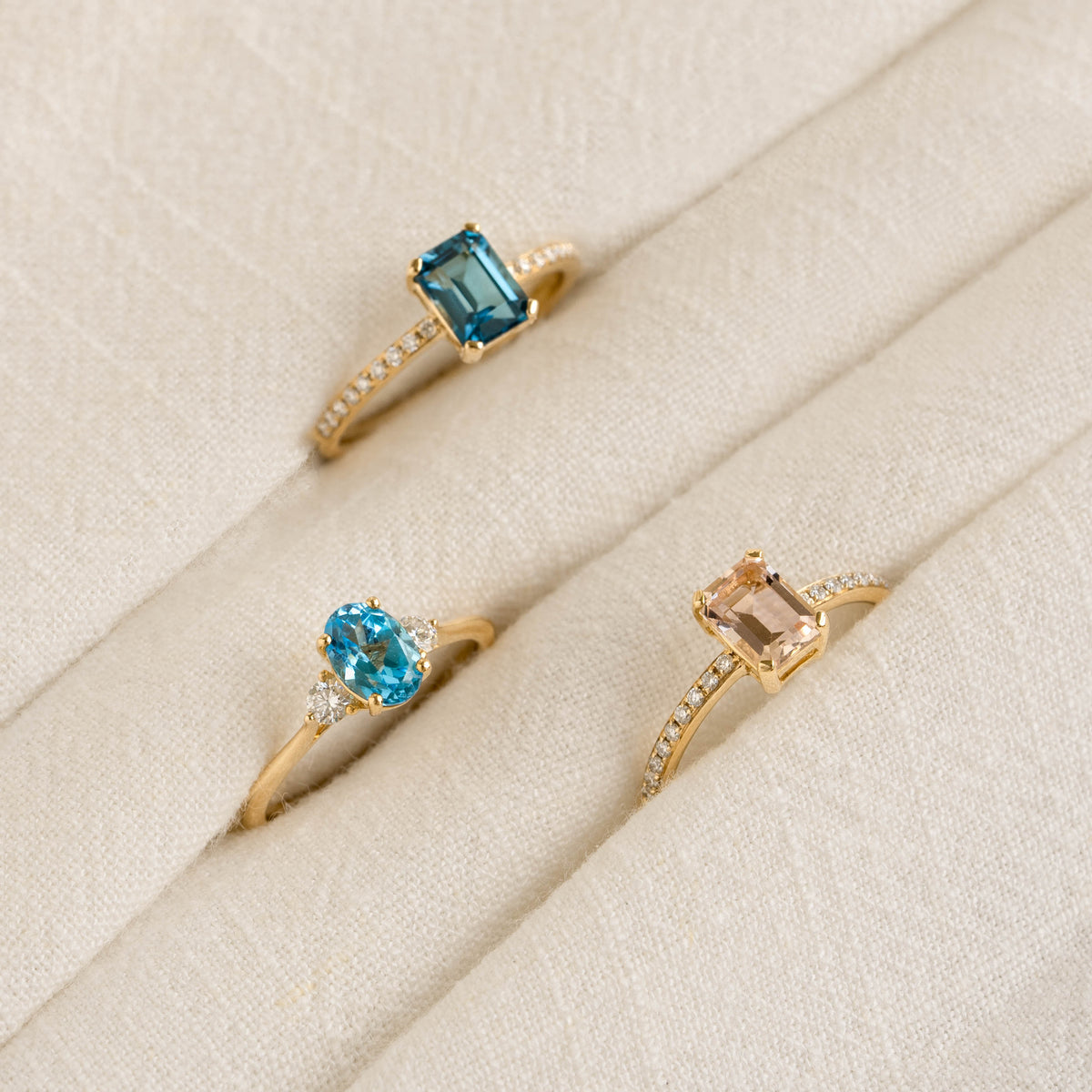 Three gold rings with blue and peach gemstones and pavé accents displayed on a beige fabric, Baguette Morganite and Diamond Ring 14k Gold - Safiya