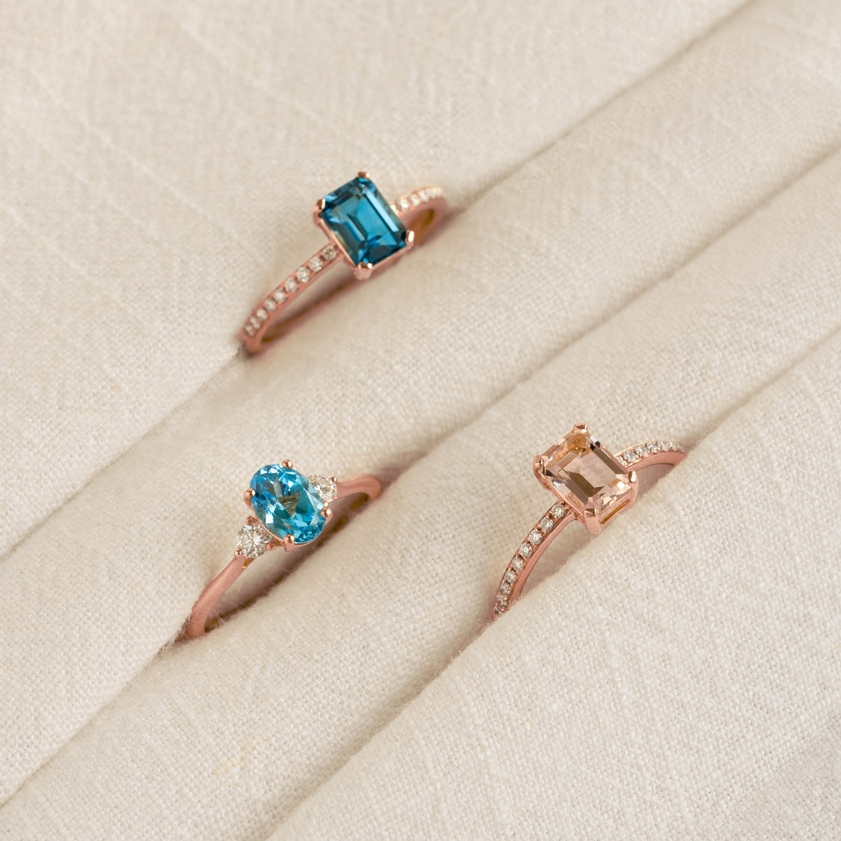 Three rose gold rings with baguette london blue topaz and diamond stones on a beige fabric display for Baguette London Blue Topaz and Diamond Ring Rose Gold - Safiya