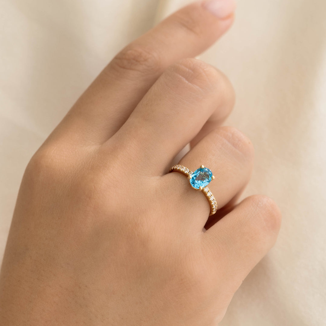 Hand wearing Oval Swiss Blue Topaz and Diamond Ring 14k Gold - Minette with pavé diamonds on a soft beige background