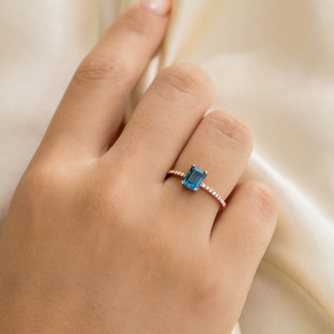Close-up of a hand wearing Baguette London Blue Topaz and Diamond Ring Rose Gold - Safiya on a cream satin background