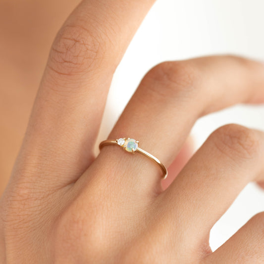 Close-up of a hand wearing the Opal and Diamond Ring - Arete with a gold band on a white background