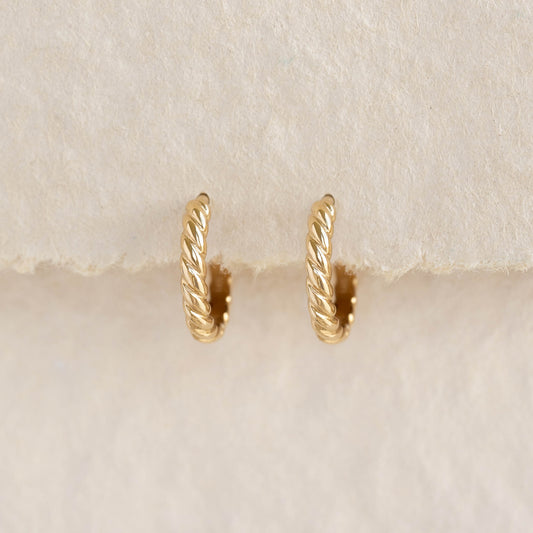 Close-up of Twist Huggie Earrings 14k Gold - Clemence with braided design on a soft beige fabric background