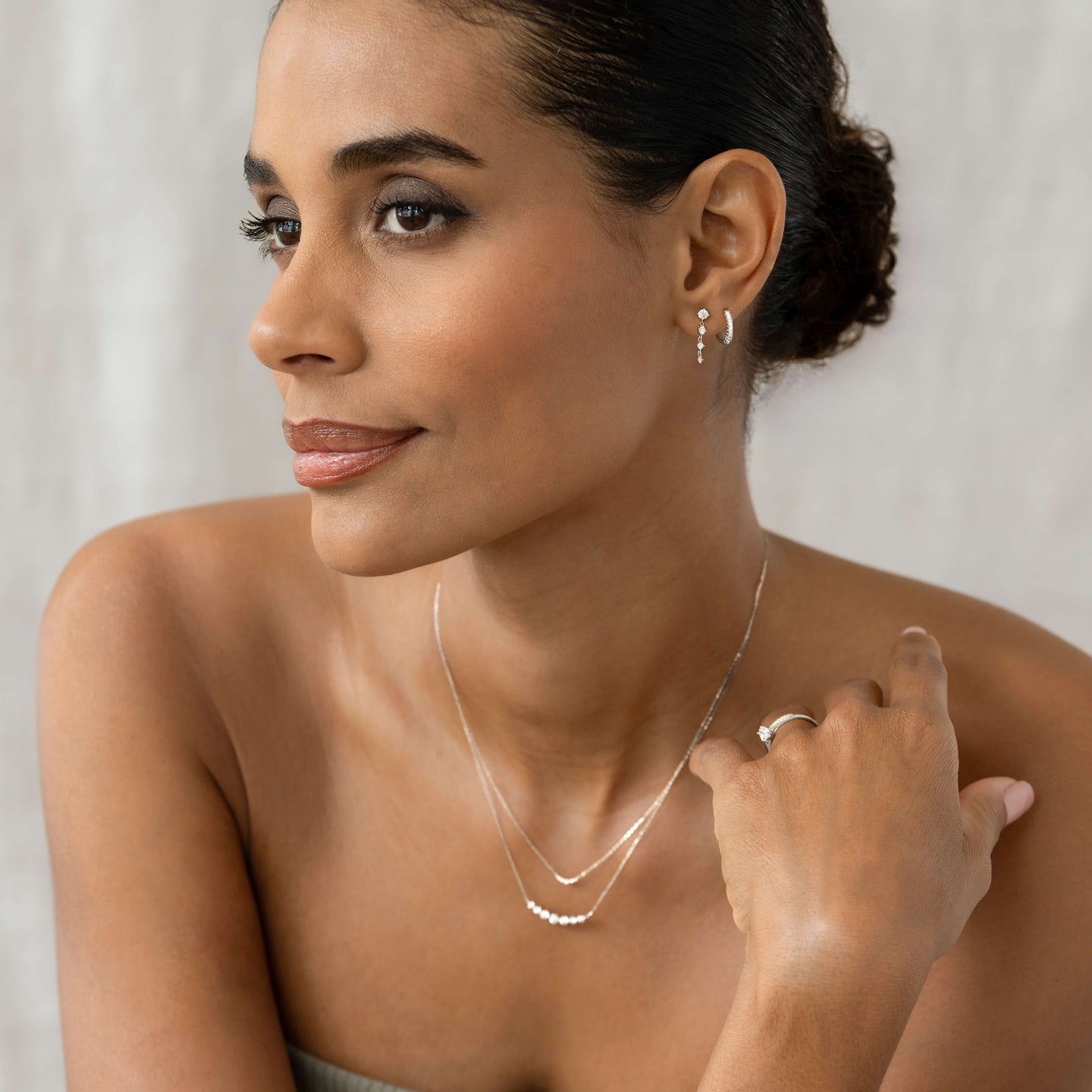 Woman wearing 14k White Gold Diamond Huggie Earrings - Susanna with layered delicate necklaces and a ring on light background
