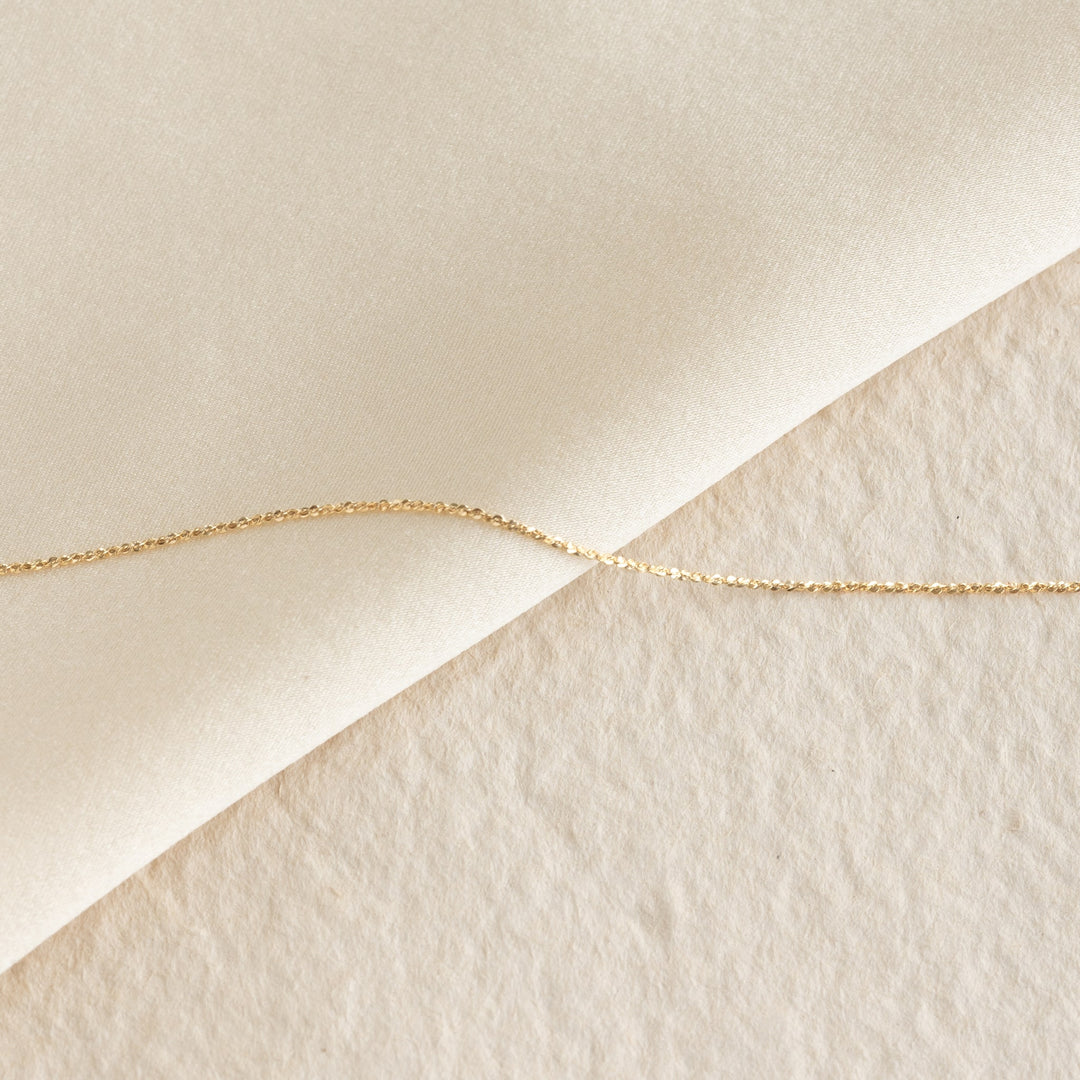 Close-up of the Tinsel Chain Necklace 14k Gold - Embla laid diagonally on soft beige textured fabric background