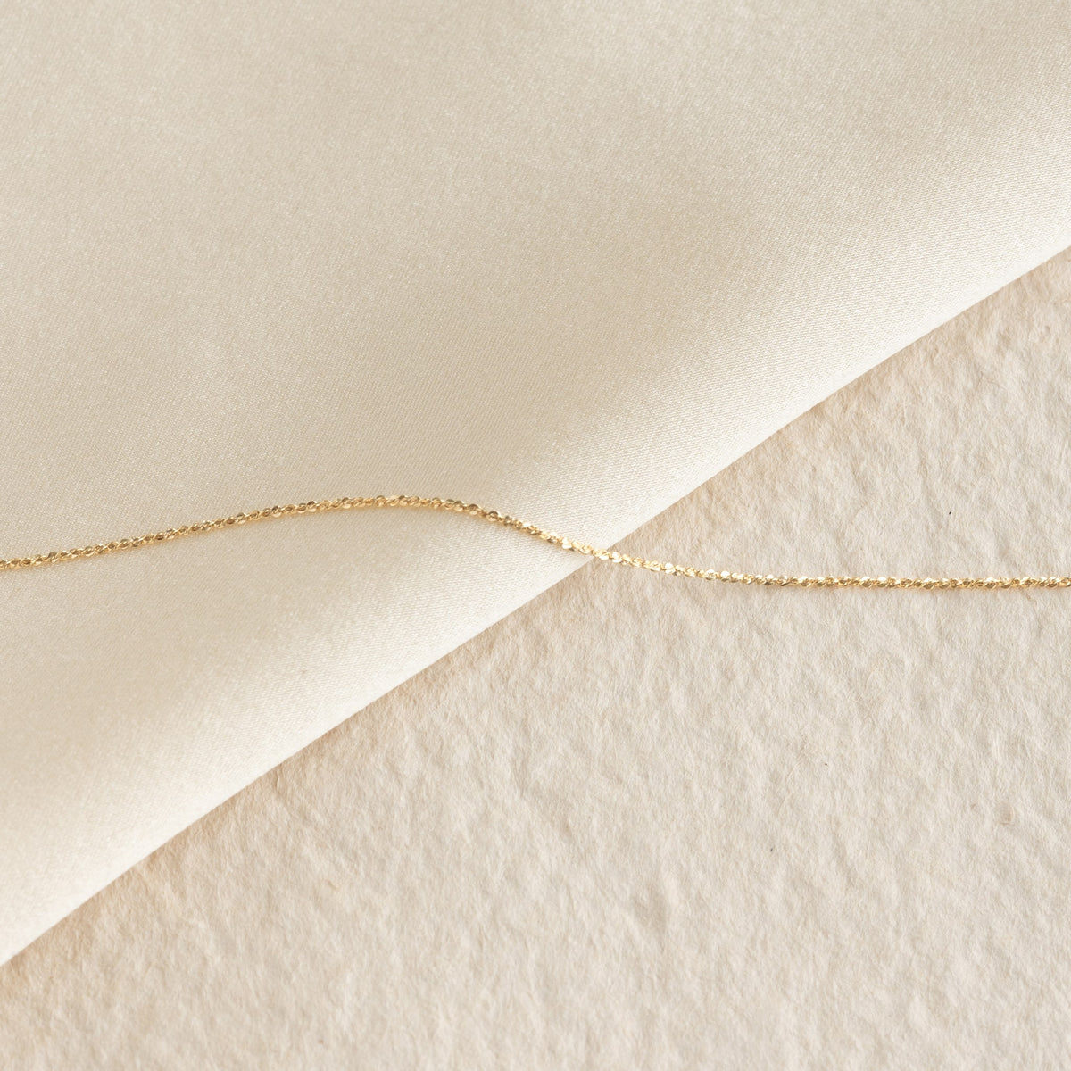 Close-up of the Tinsel Chain Necklace 14k Gold - Embla laid diagonally on soft beige textured fabric background