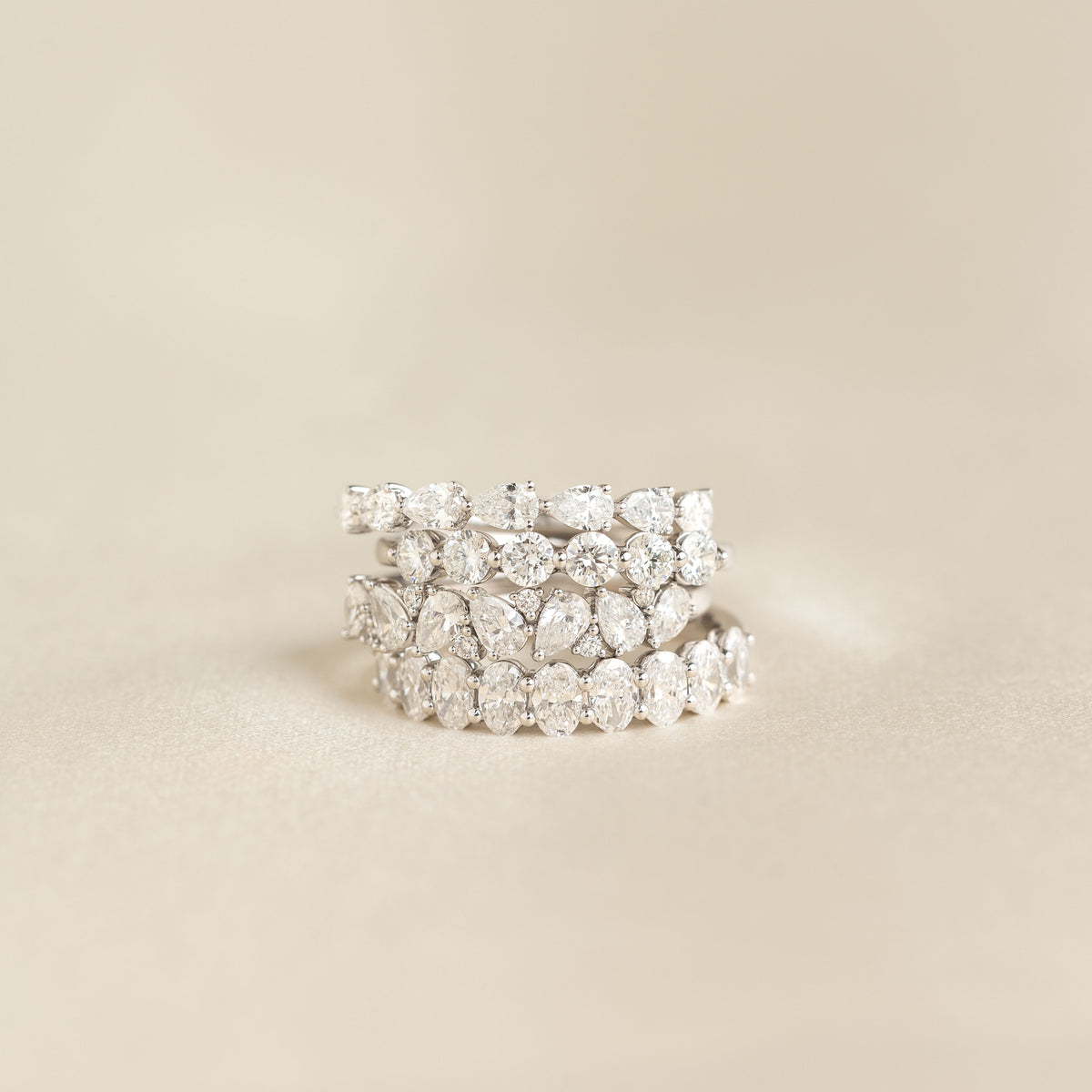 A stack of four White Gold rings set with diamonds, featuring alternating pear-shaped and round stones, arranged in a layered style against a soft beige background.