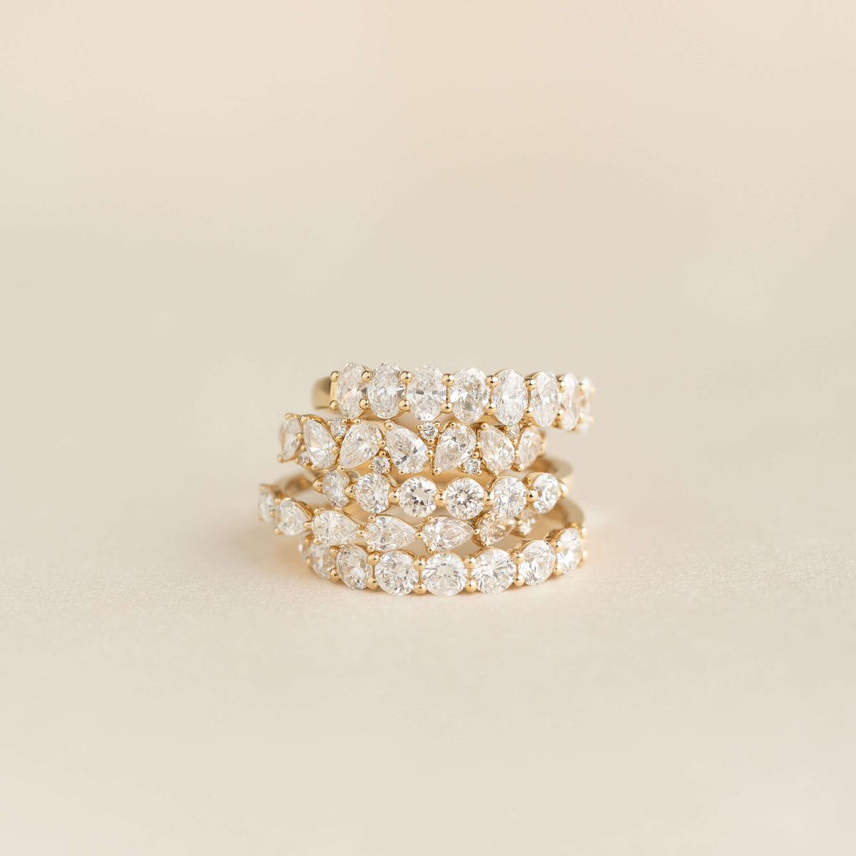 A stack of Yellow Gold rings set with diamonds, featuring alternating pear-shaped and round stones, arranged in layered bands against a soft beige background.