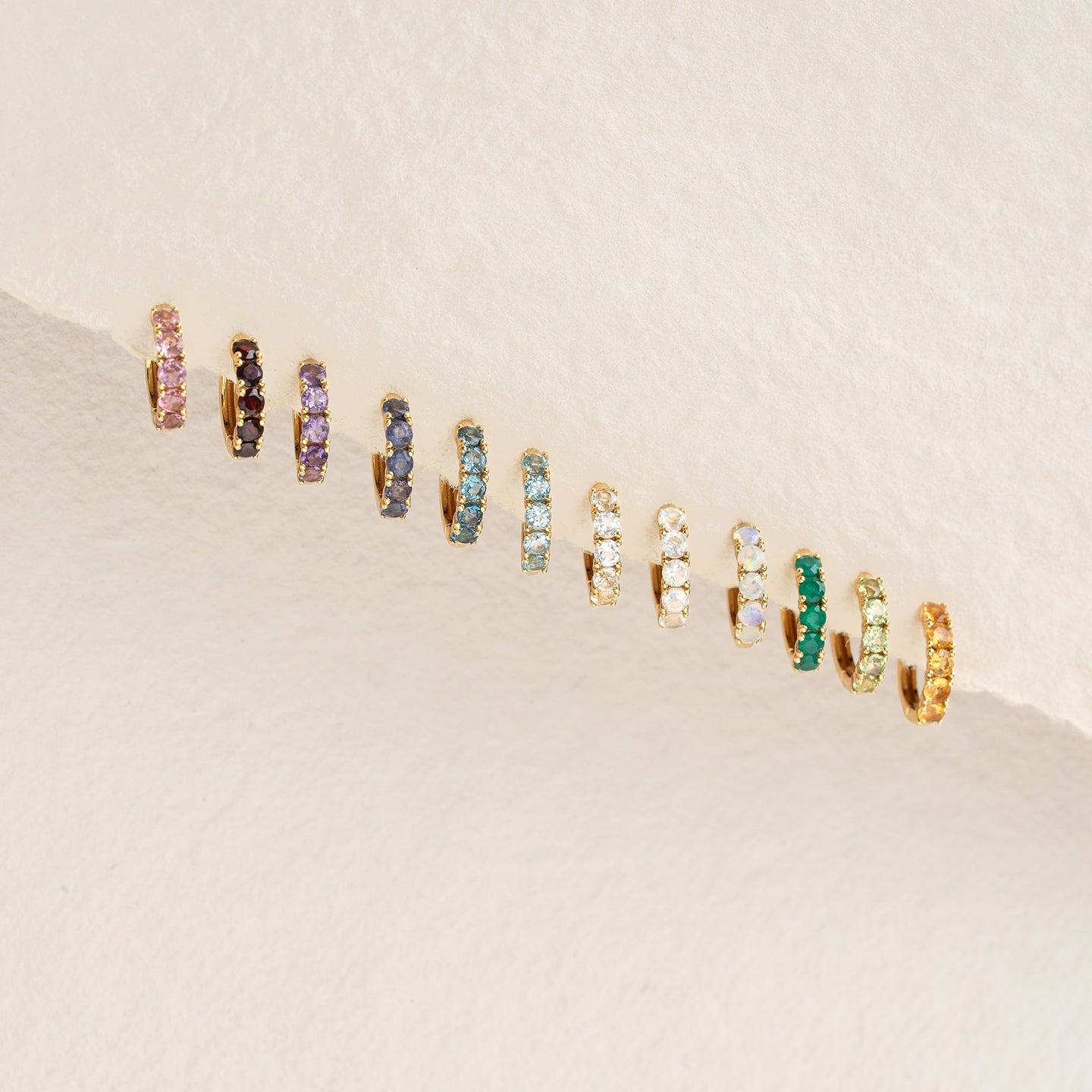 Row of colorful gemstone huggie earrings including the October Birthstone Huggie Earrings - Opal on a neutral background