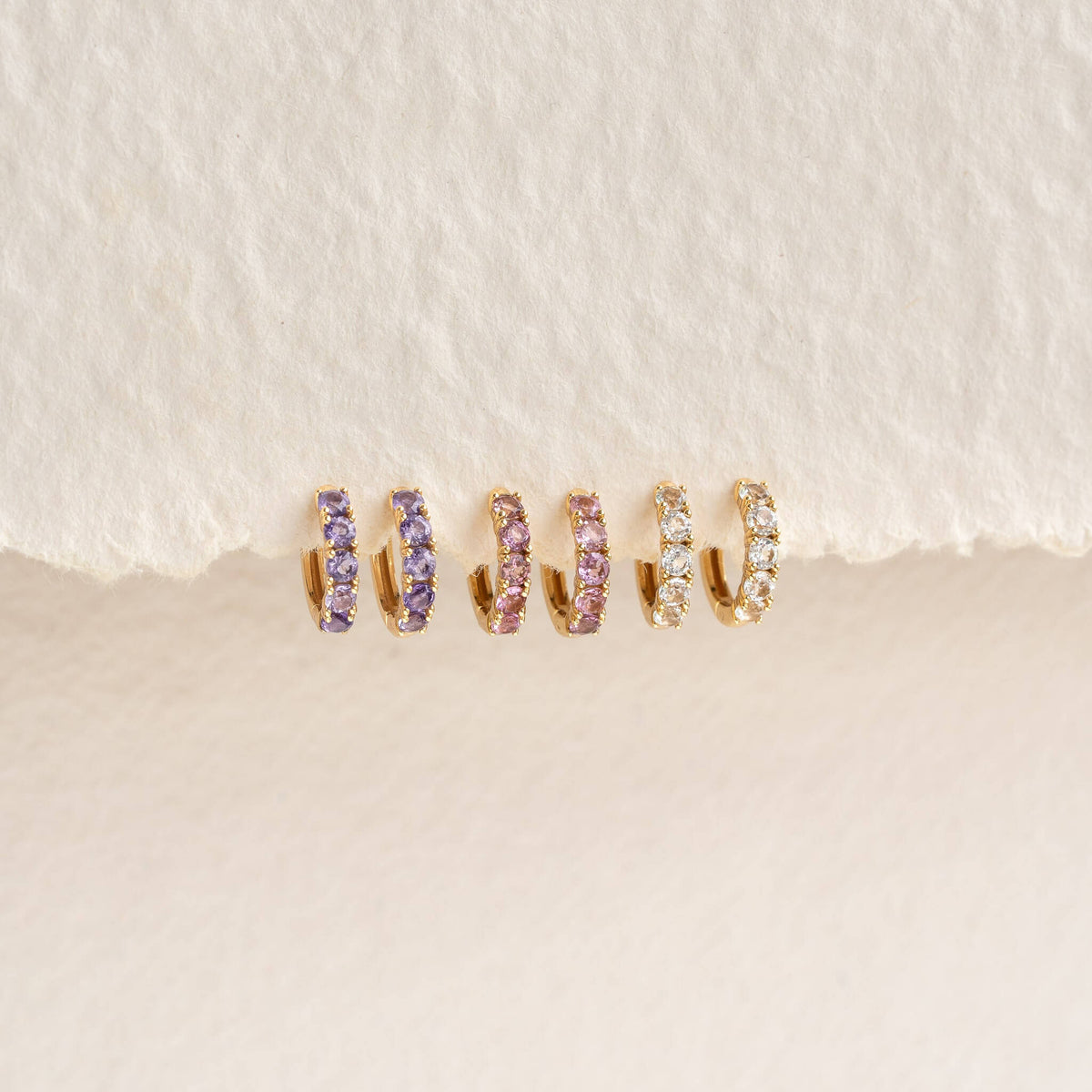 Three pairs of February Birthstone Huggie Earrings - Amethyst in gold vermeil with purple and clear gemstones on textured background