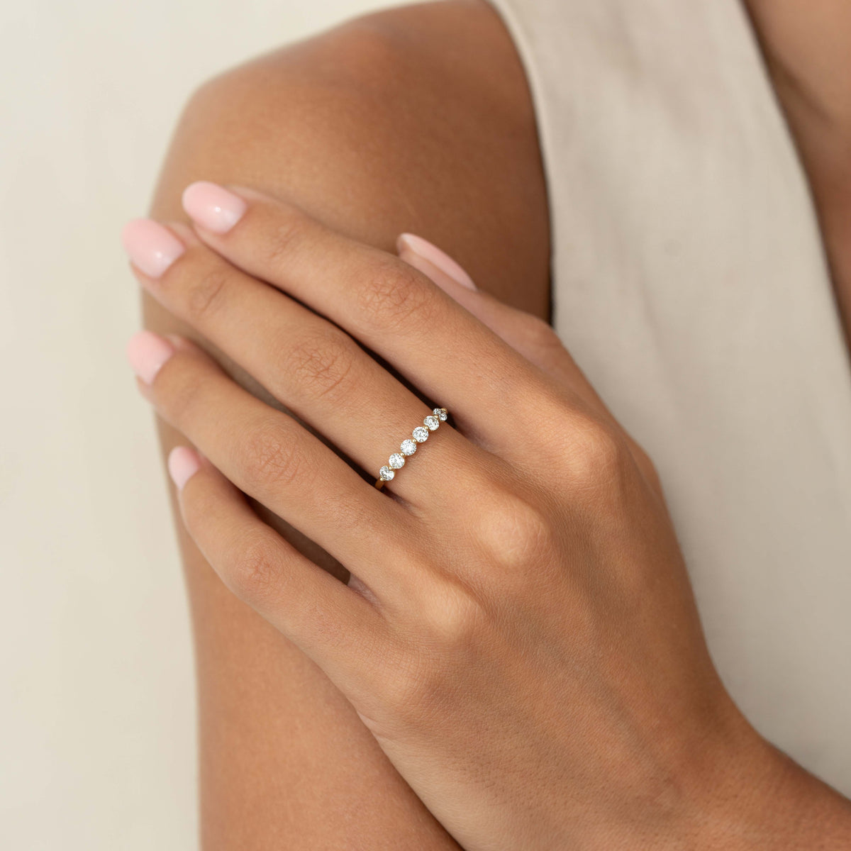 A close-up of a hand resting on a shoulder, wearing a delicate Yellow Gold ring set with a row of round diamonds, against a soft neutral background.