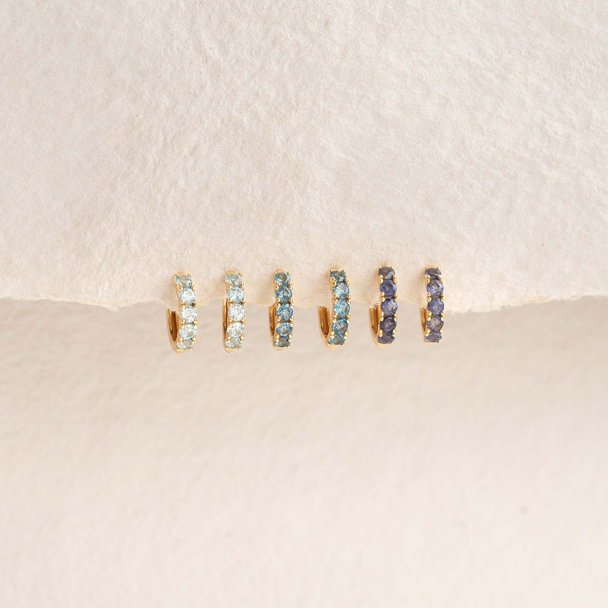 Three pairs of March Birthstone Huggie Earrings - Blue Topaz featuring gold vermeil and blue gemstones on a textured background