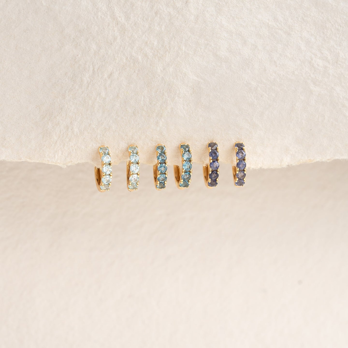 September Birthstone Huggie Earrings - London Blue Topaz in gold vermeil with varying blue gemstones on a cream background