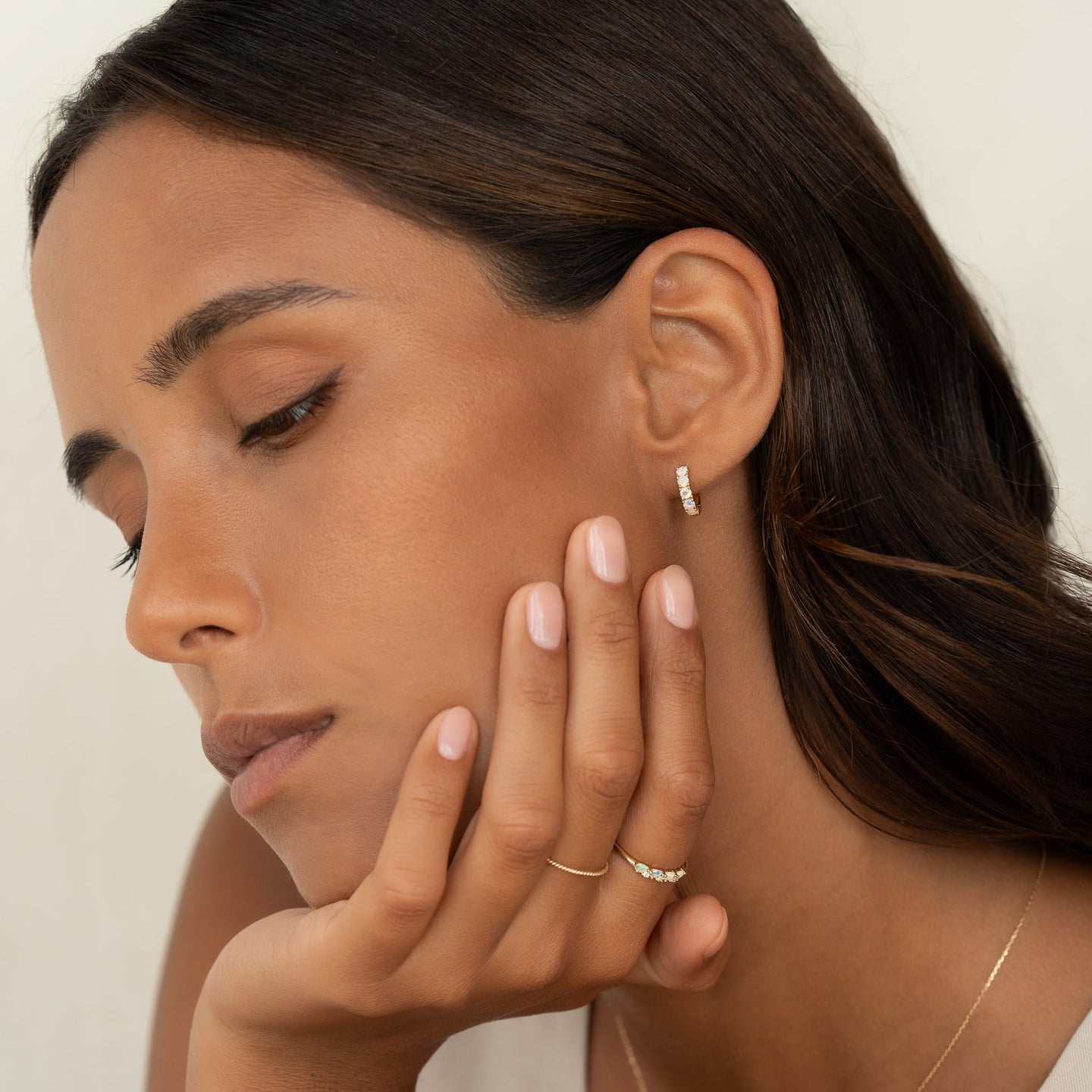 Close-up of a woman wearing October Birthstone Huggie Earrings - Opal with gold vermeil setting and natural makeup on a cream background