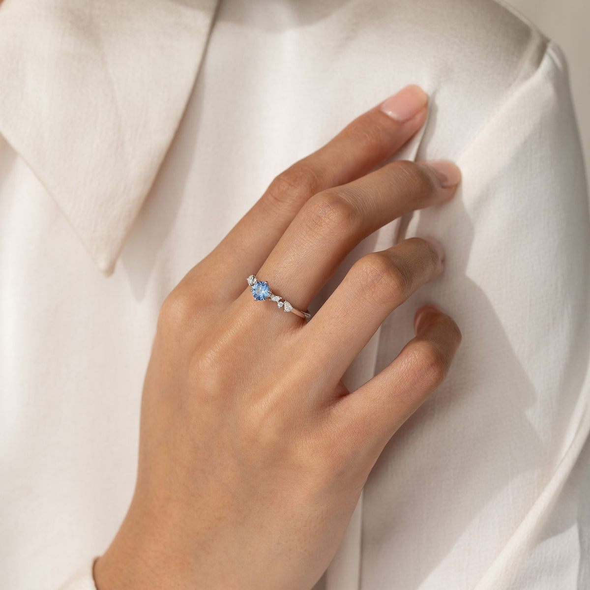 On her middle finger, she wears a delicate White Gold ring featuring a vibrant, round brilliant-cut blue sapphire center stone flanked by an asymmetrical cluster of small white diamonds.