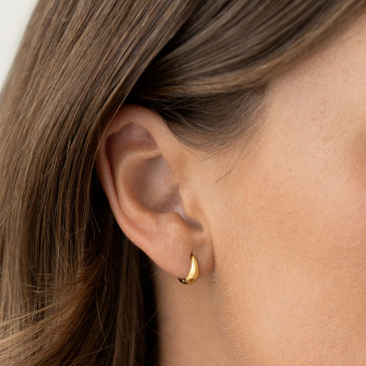 Close-up of a woman wearing Dome Huggie Earrings - Cassia in gold vermeil on her ear against a neutral background