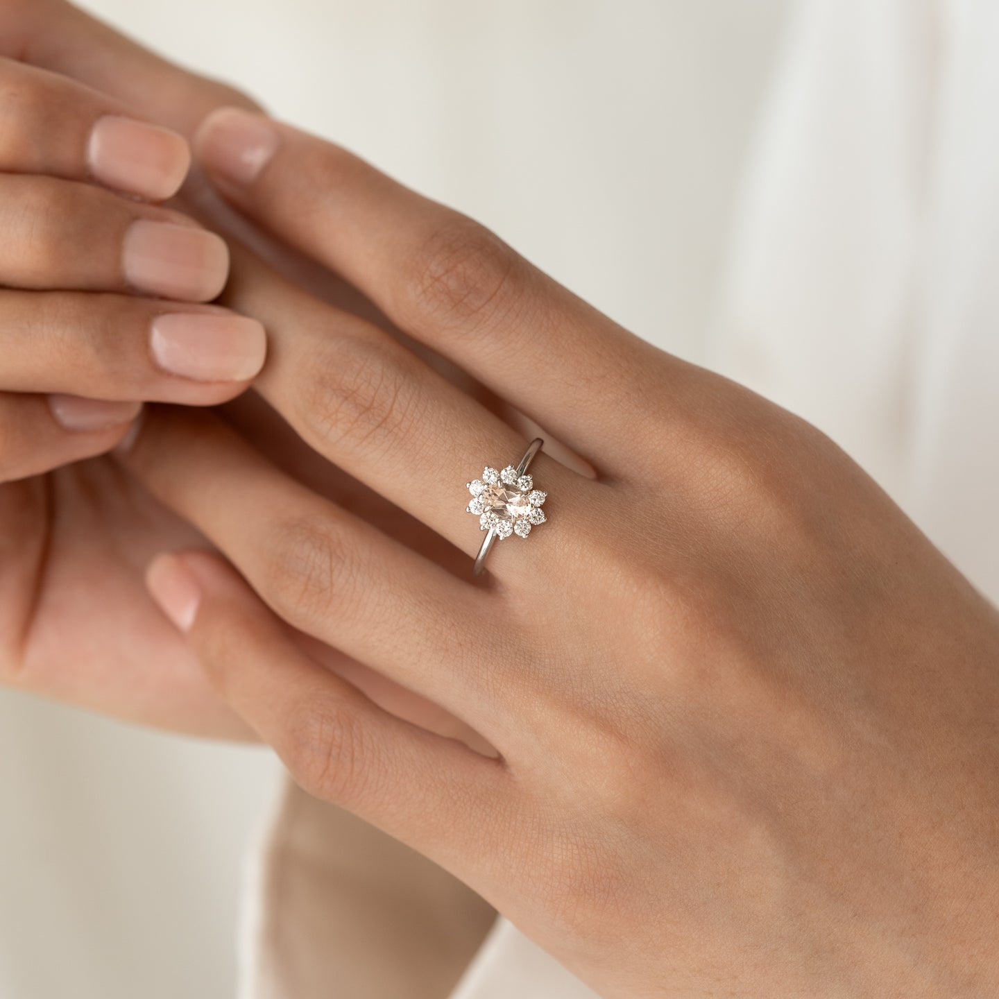 Close-up of hand wearing Sunburst Morganite Halo Ring White Gold - Evelina with oval gemstone and diamond halo on neutral background