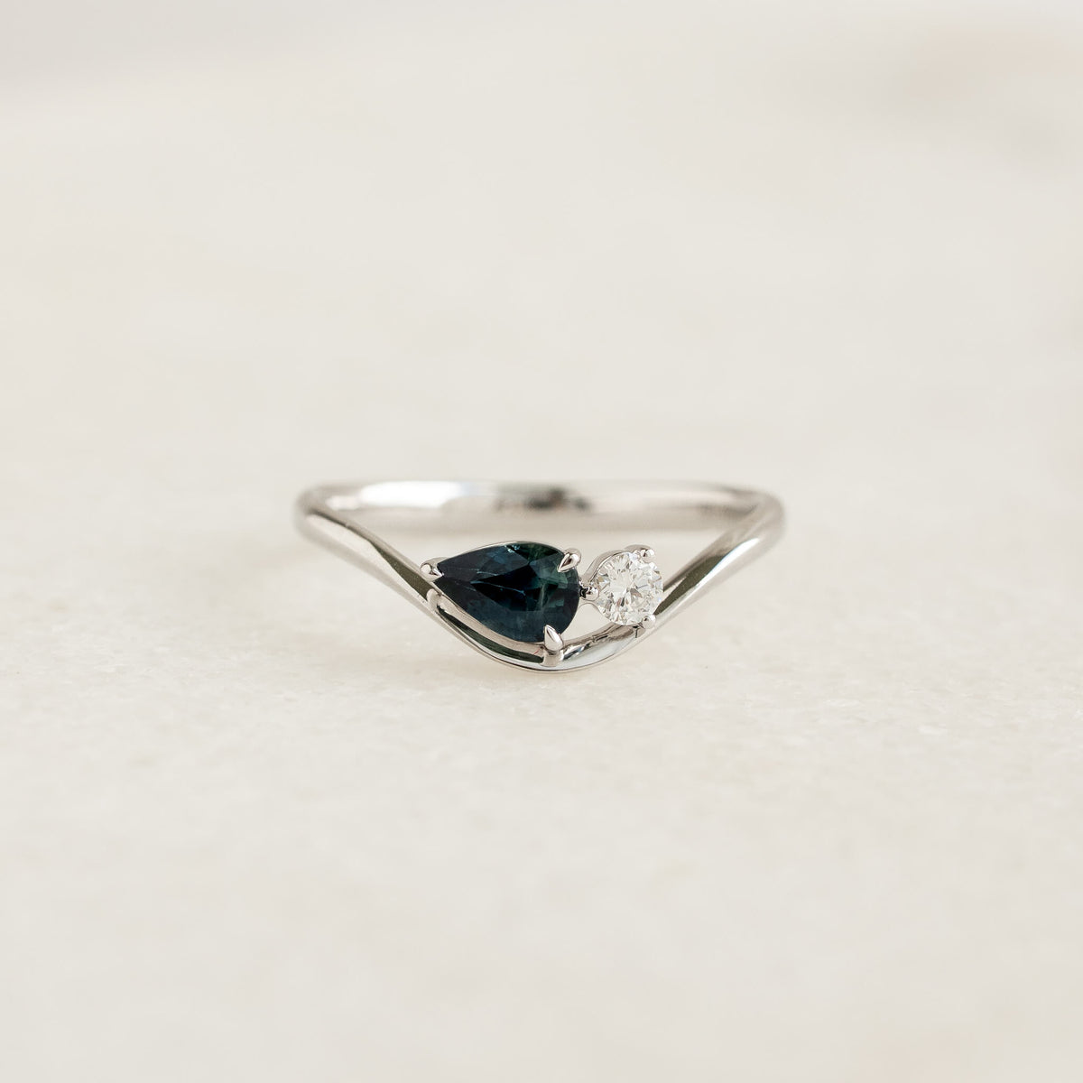 A close-up shot of the Lekha pear sapphire ring crafted in 14k White Gold. The ring features a gentle, asymmetrical wavy band that curves downward to cradle a pear-cut, deep teal-blue sapphire set horizontally.