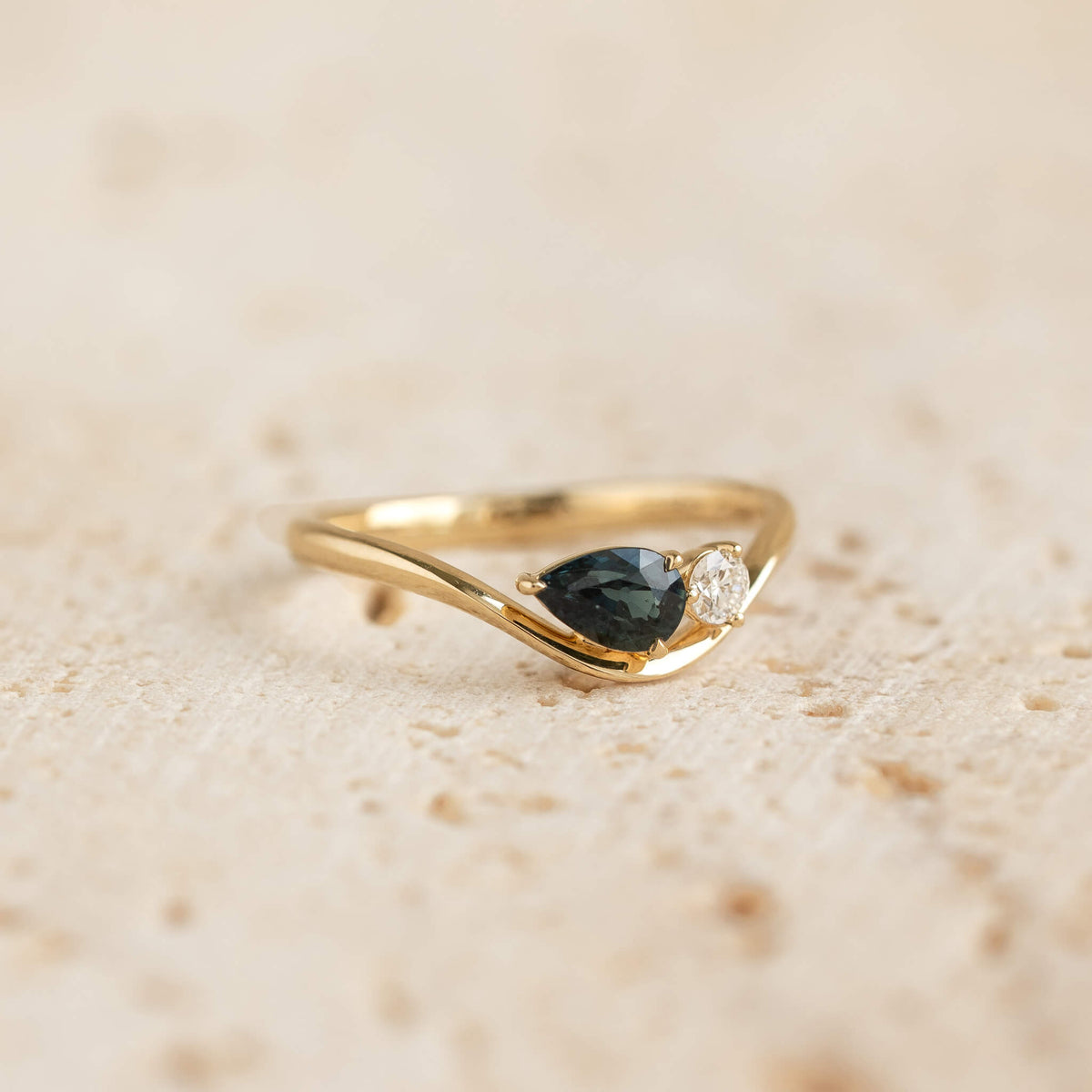 A close-up shot of the Yellow Gold pear sapphire ring crafted in 14k yellow gold. The ring features a gentle, asymmetrical wavy band that curves downward to cradle a pear-cut, deep teal-blue sapphire set horizontally.