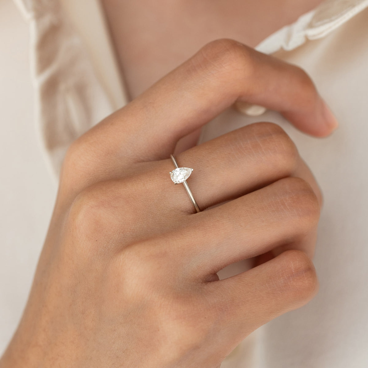 A hand wearing a delicate White Gold ring with a pear-shaped diamond set at the center, resting on light, smooth skin. The background is a soft cream fabric.