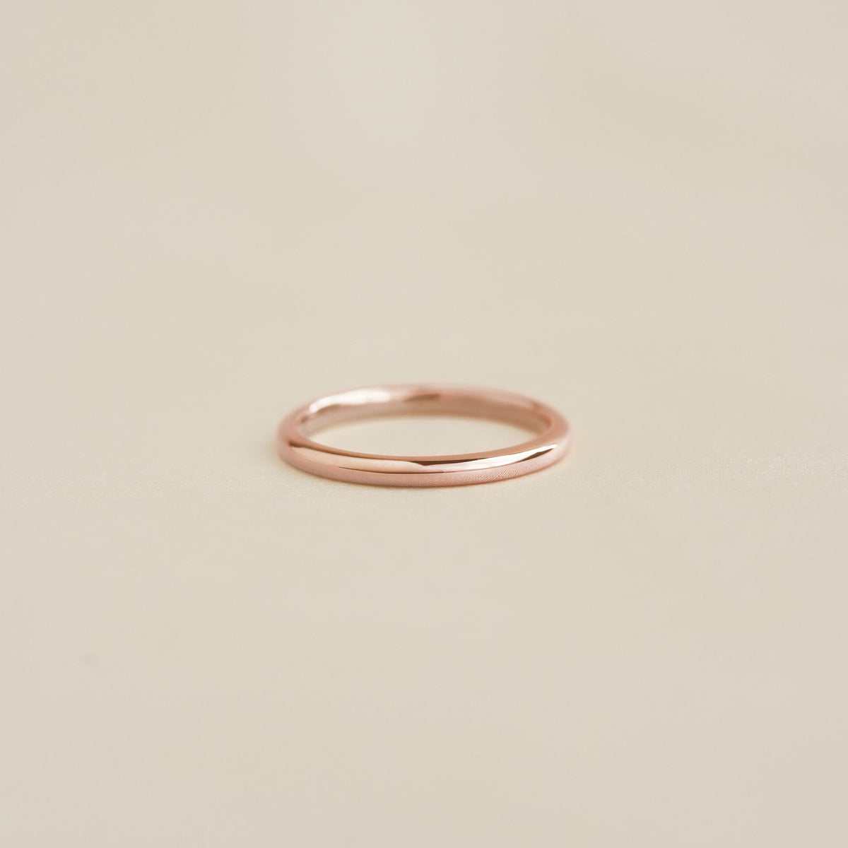 Close-up of a slim polished Round Rose Gold Band - Marni resting on a soft beige surface with subtle reflections