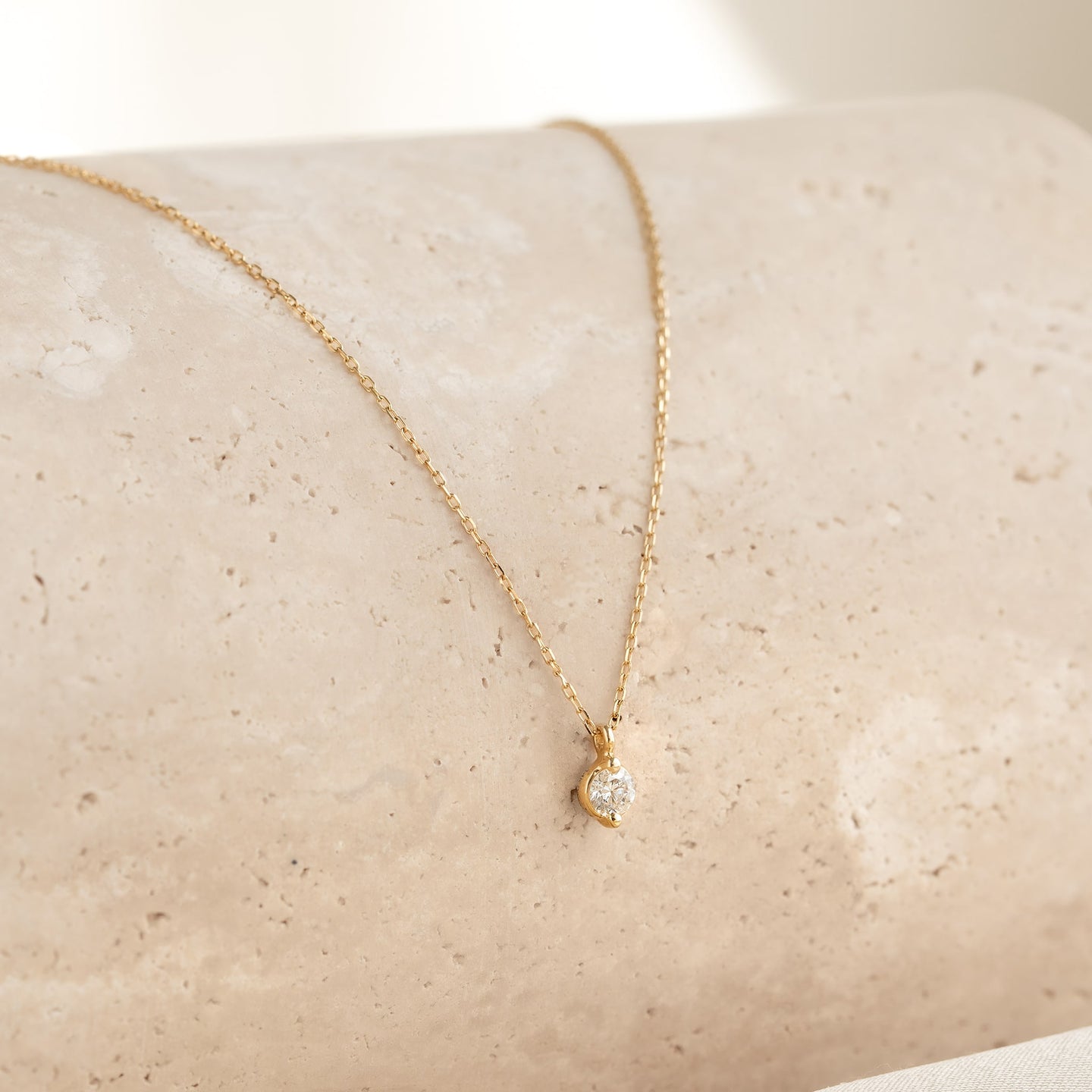 Close-up of a delicate Diamond Pendant Necklace 14k Gold - Atiana resting on a beige textured surface with soft natural light